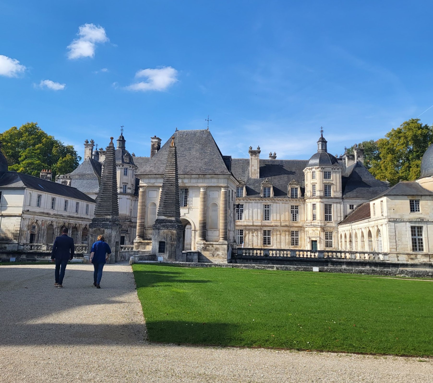 Deux personnes marchent sur une allée de gravier devant le château de Tanlay (Yonne 89) aux toits gris et murs clairs, sous un ciel bleu avec quelques nuages blancs.