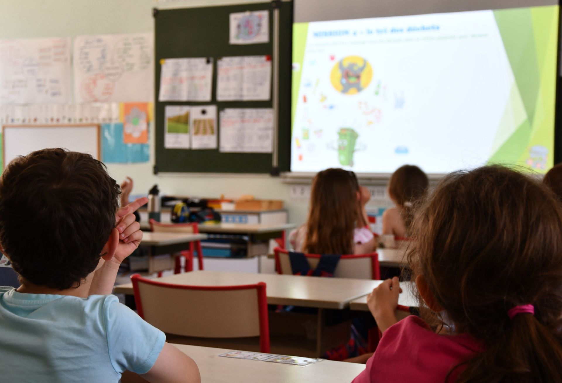 Des écoliers regardent un tableau numérique projeté dans une salle de classe. Un enfant lève la main.