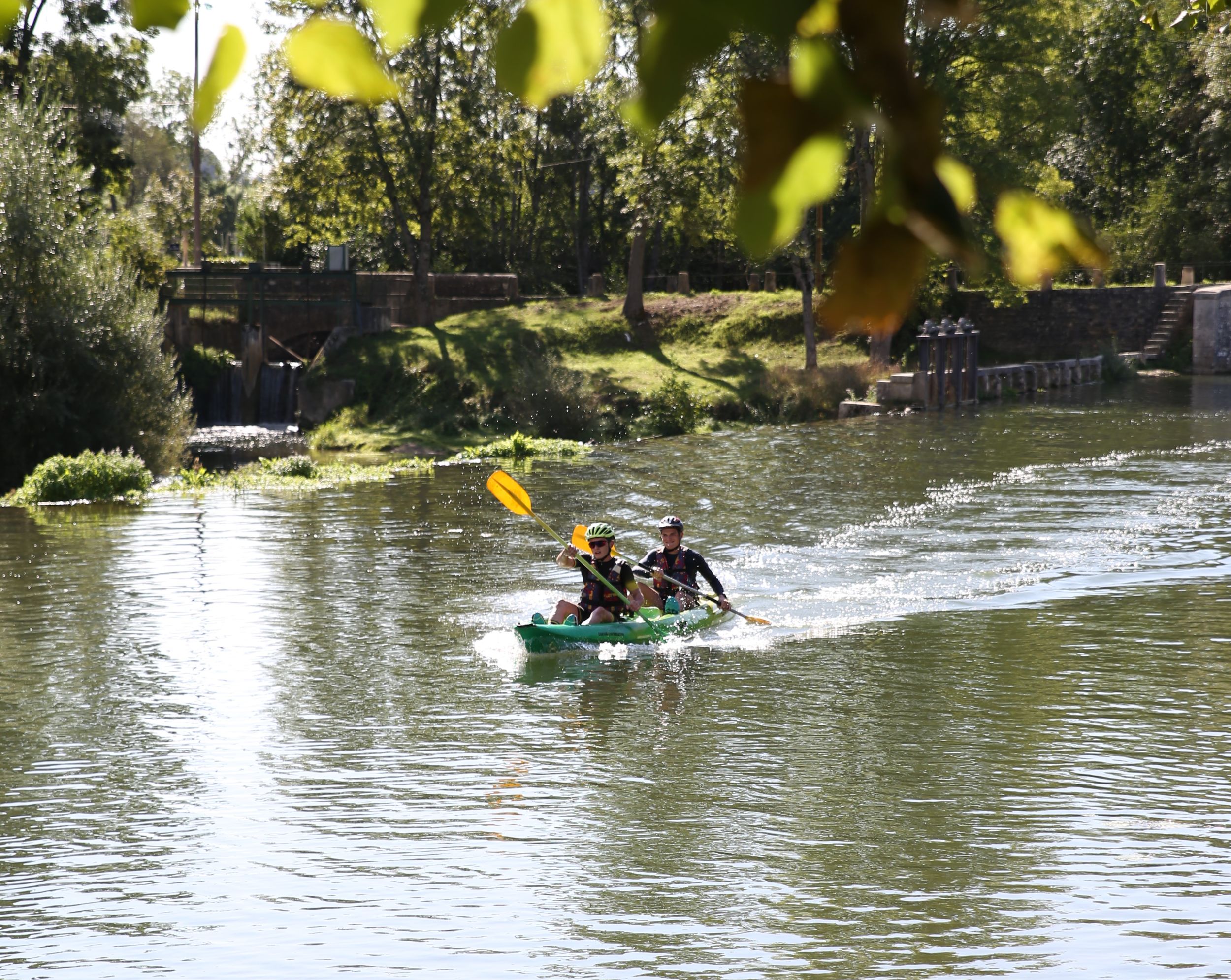 Deux personnes en kayak biplace naviguent sur une rivière calme, entourées d'arbres luxuriants et d'une structure en pierre au loin.
