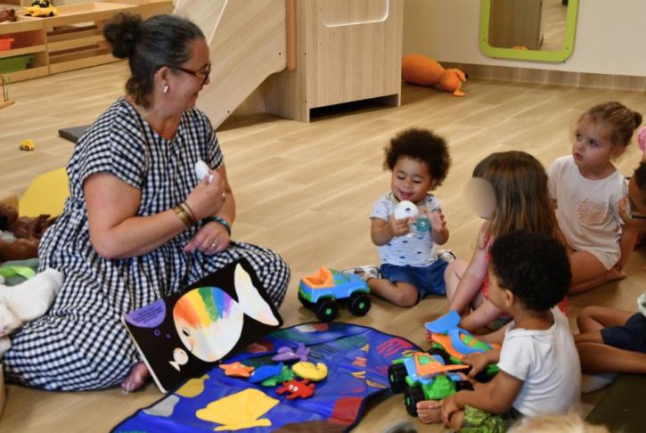 Une éducatrice assise par terre montre un livre illustré d'un poisson arc-en-ciel à des enfants attentifs. Des jouets sont dispersés autour d'eux.