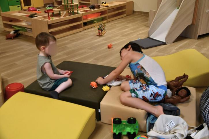 Deux enfants jouant avec des voitures miniatures et des poupées sur des coussins dans une salle de jeux aménagée.